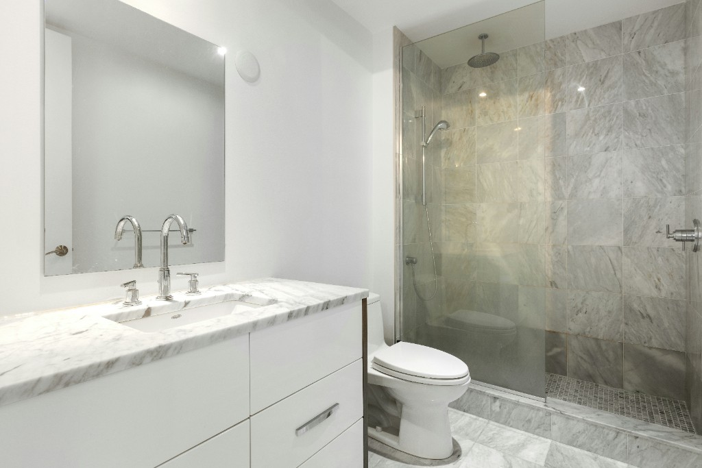 Baño moderno con mueble blanco de mármol, grifería cromada, espejo amplio y ducha acristalada con revestimiento gris.