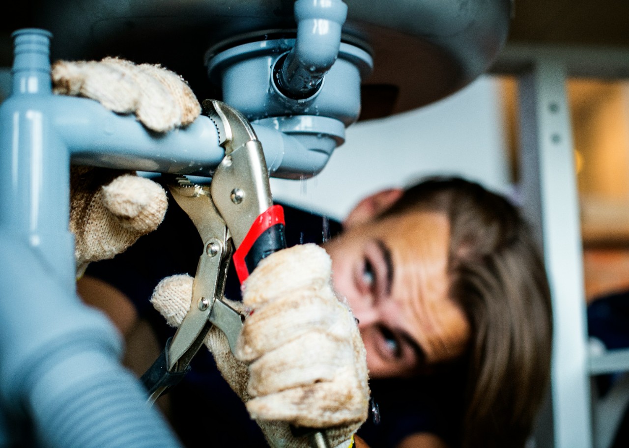 Mujer con guantes beige usando alicates para reparar tubería azul bajo el fregadero. Imagen en primer plano de trabajo de fontanería.