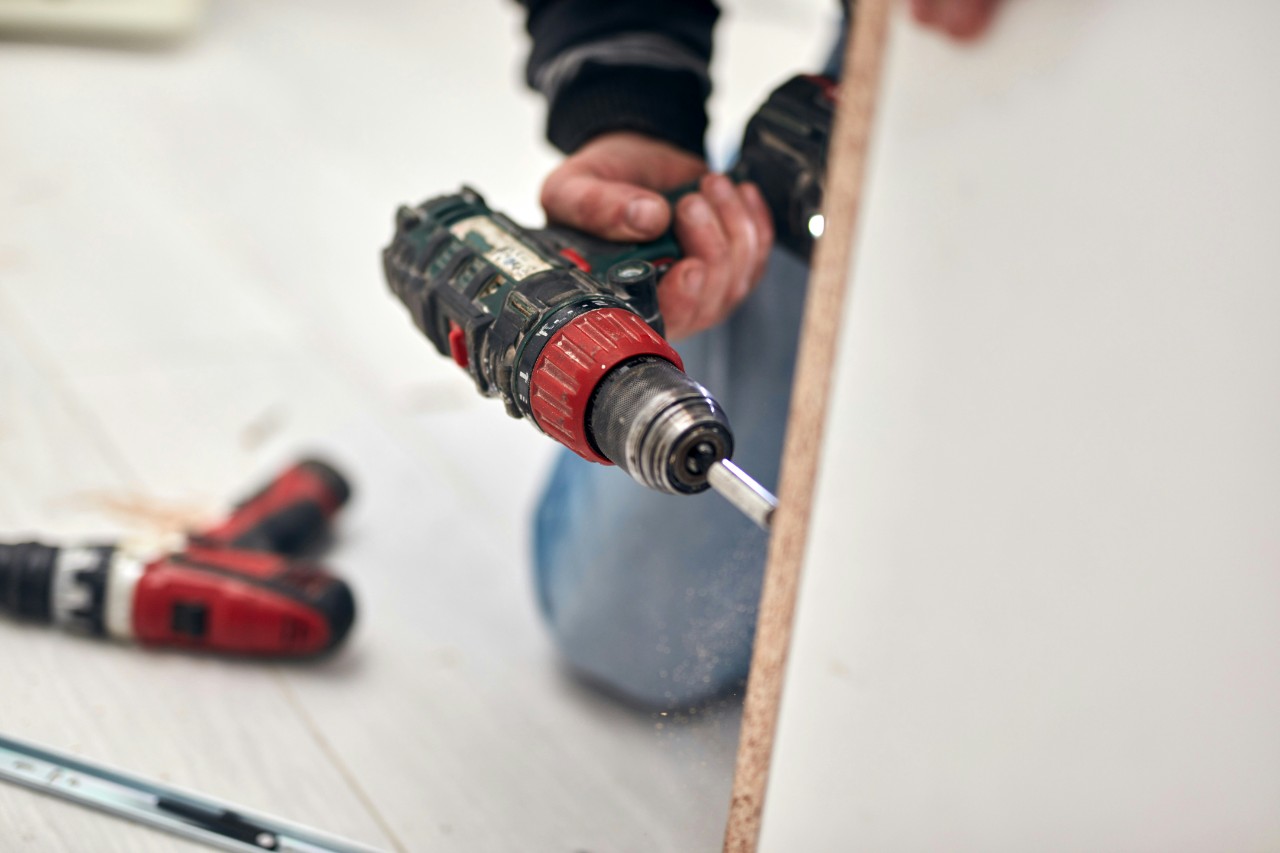 Persona sosteniendo un taladro atornillador negro con detalles rojos apuntando hacia una superficie de madera clara.