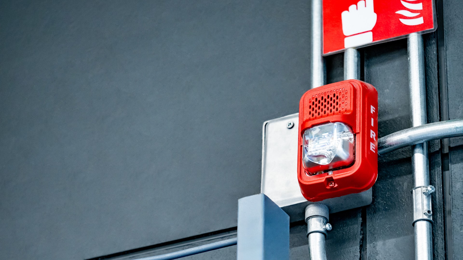 Alarma de incendios roja instalada en pared gris junto a tubería metálica con señal de extintor visible arriba