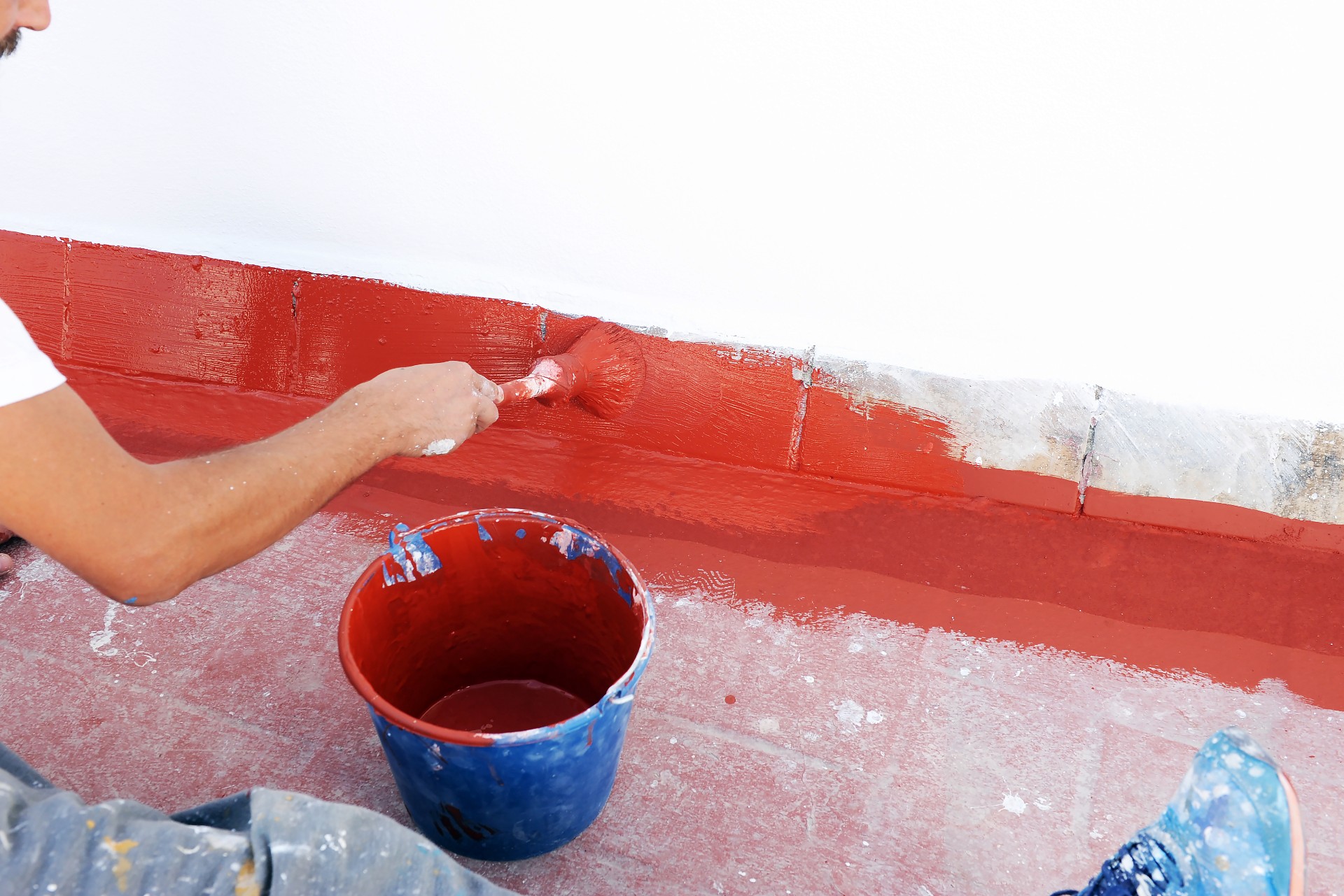 Persona pintando una pared exterior de color rojo con brocha, cubo azul de pintura en el suelo sobre superficie manchada