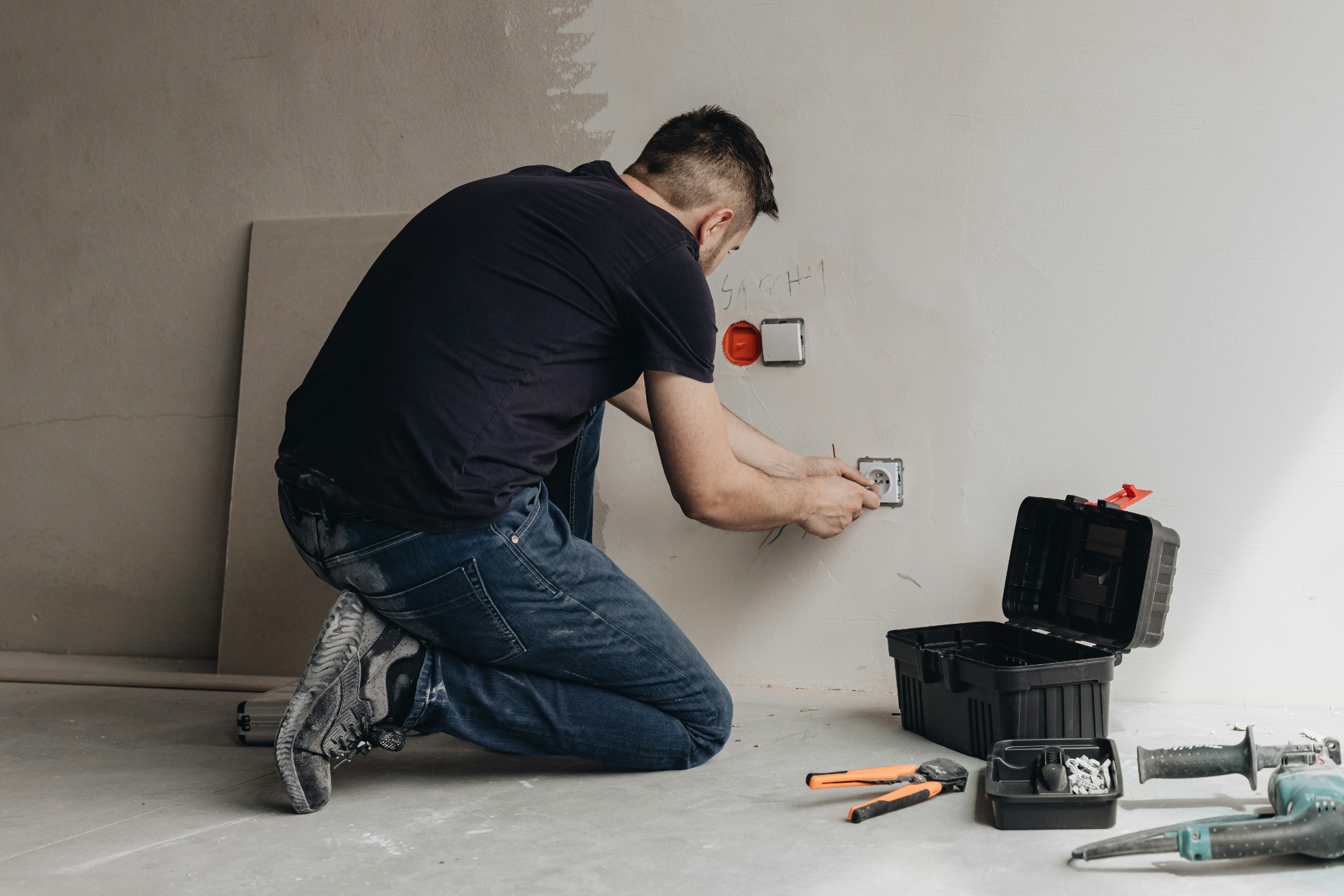 Electricista instalando enchufes en pared blanca, arrodillado con herramientas y caja de trabajo negra en el suelo